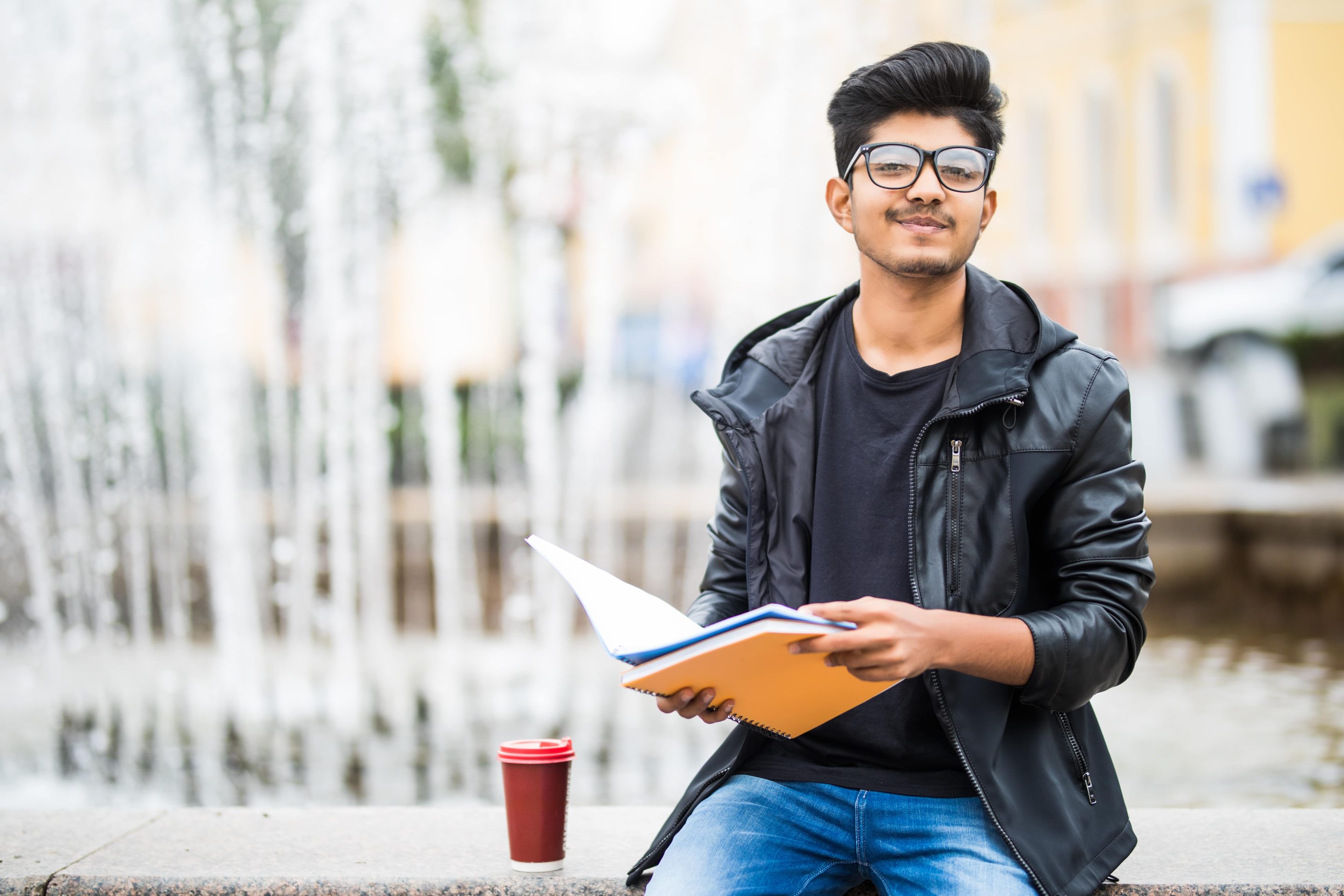 indian-student-man-holding-pile-books-sitting-near-fountain-street